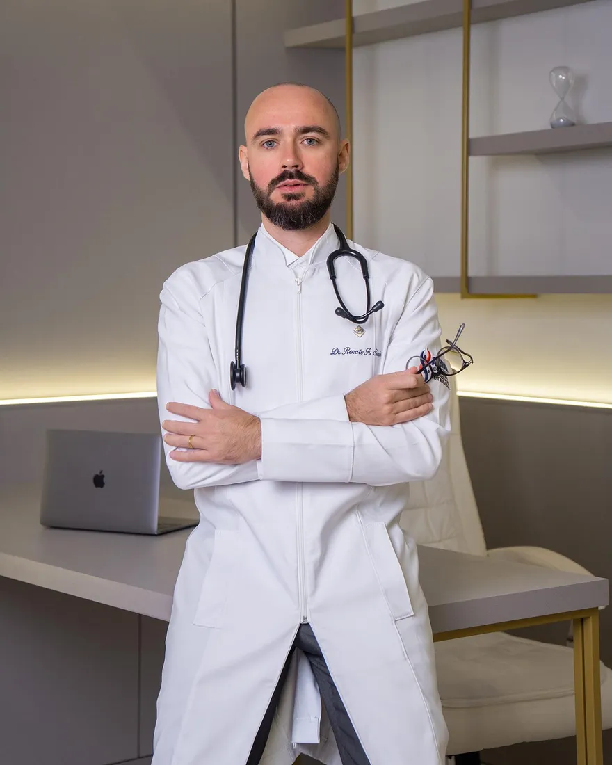 Dr. Renato Susin de jaleco branco e estetoscópio, encostado na mesa do consultório